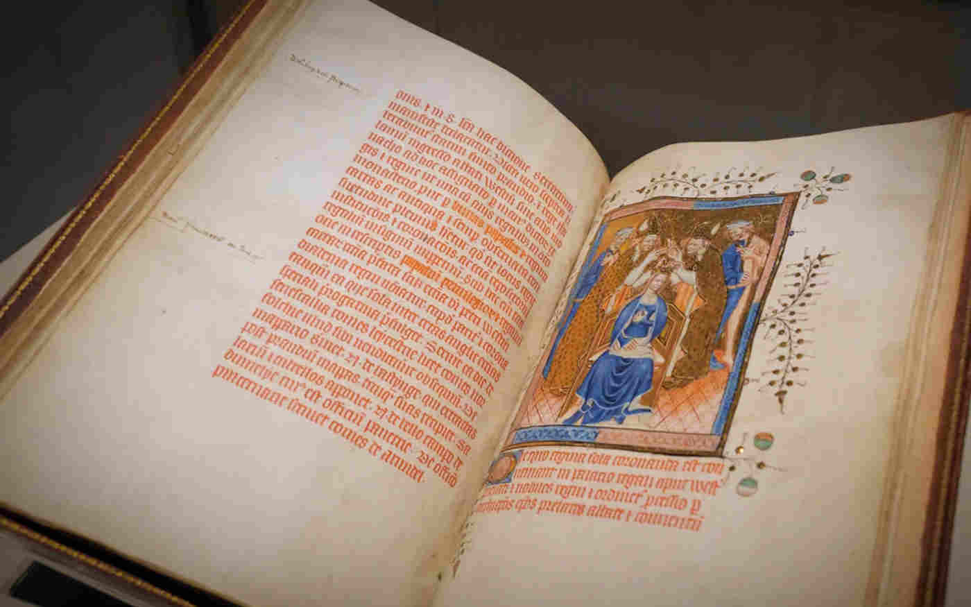Photograph of an open bound manuscript, the Liber Regalis, on display in Westminster Abbey. The left hand page contains medieval Latin text in red. The right hand page is an illumination of a queen being crowned, surrounded by members of the clergy.