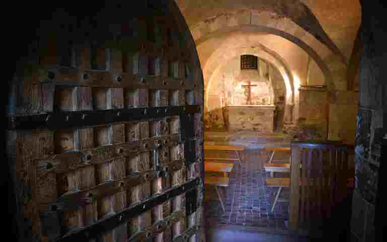 A medieval wooden door ajar revealing a small chamber with benches and a stone altar with a crucifix and candles