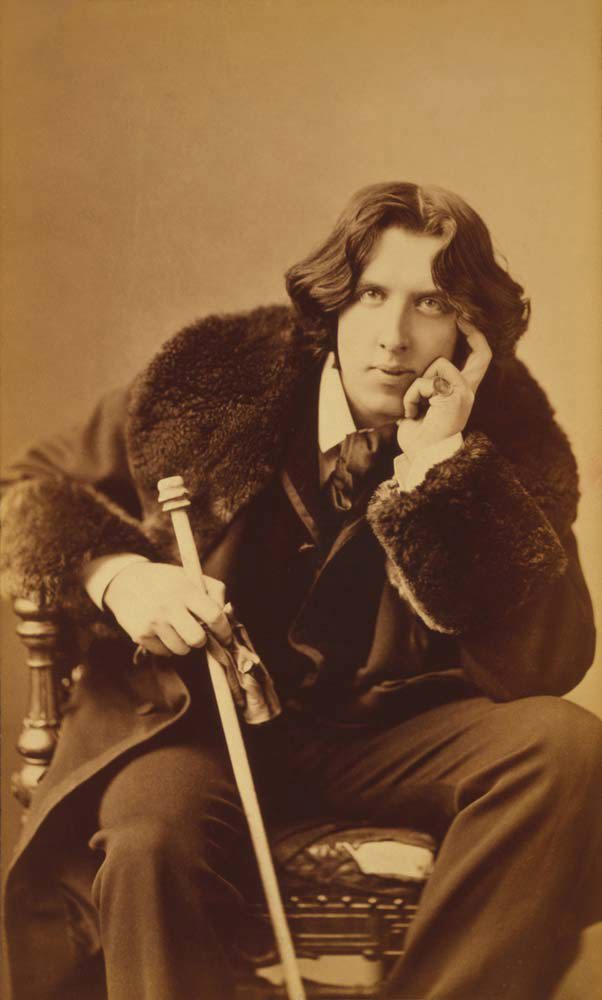 Sepia photo of Oscar Wilde, seated, wearing a fur-trimmed coat and holding a long walking stick