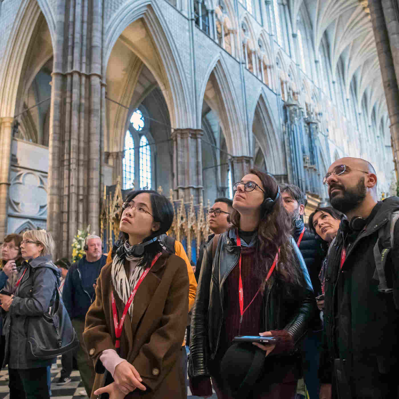 Photograph of a group of visitors looking upwards, with multimedia guides around their necks, and the stone arches above the quire visible in the background