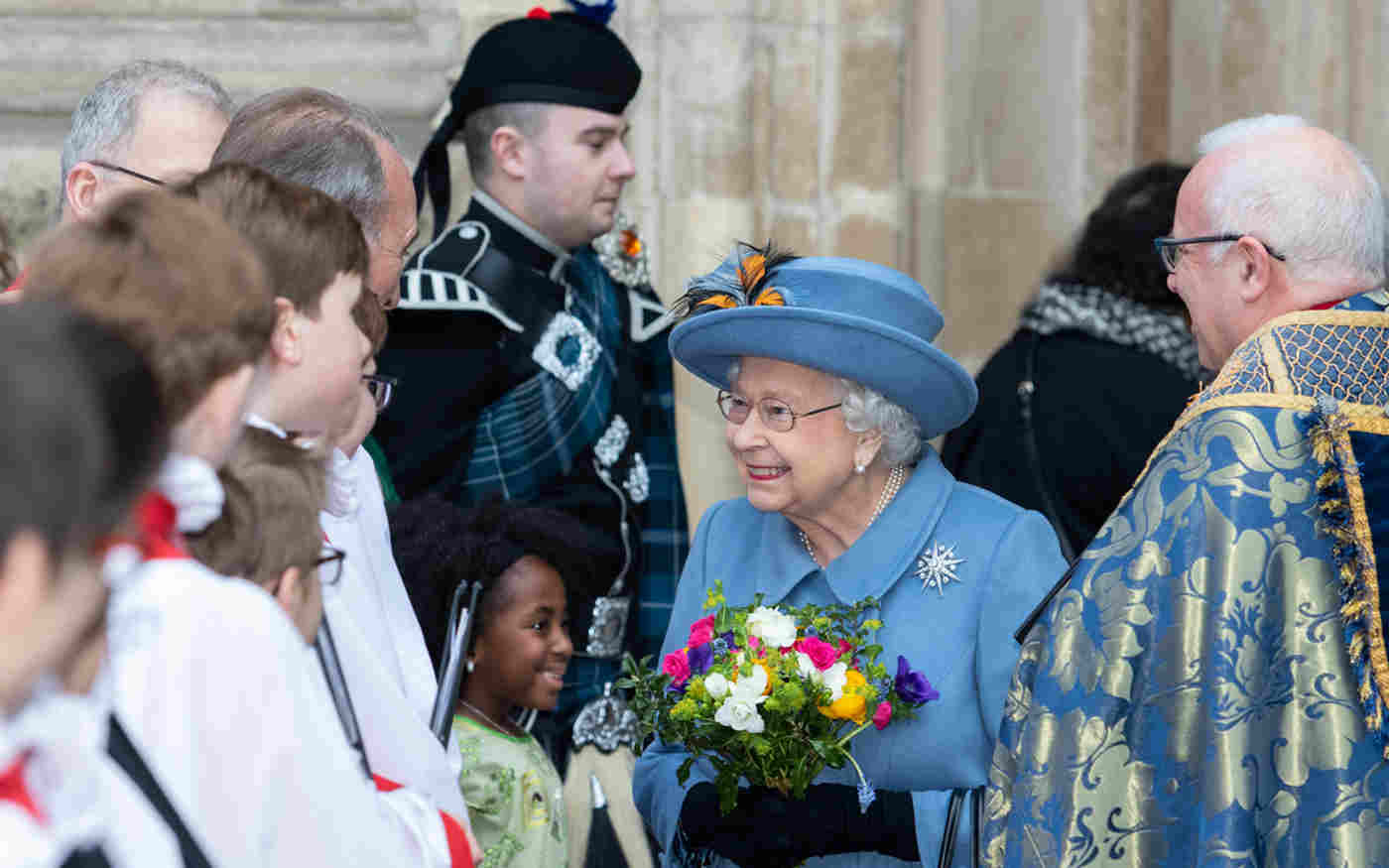 HM Queen Elizabeth II meeting choristers outside the Abbey. She is wearing a blue outfit and carrying a bouquet as she stands next to the Dean of Westminster
