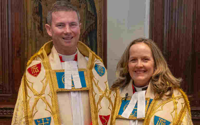 Photograph of two priests, a male and female, looking at the camera and smiling, wearing ornate clerical robes of gold, white, red and blue