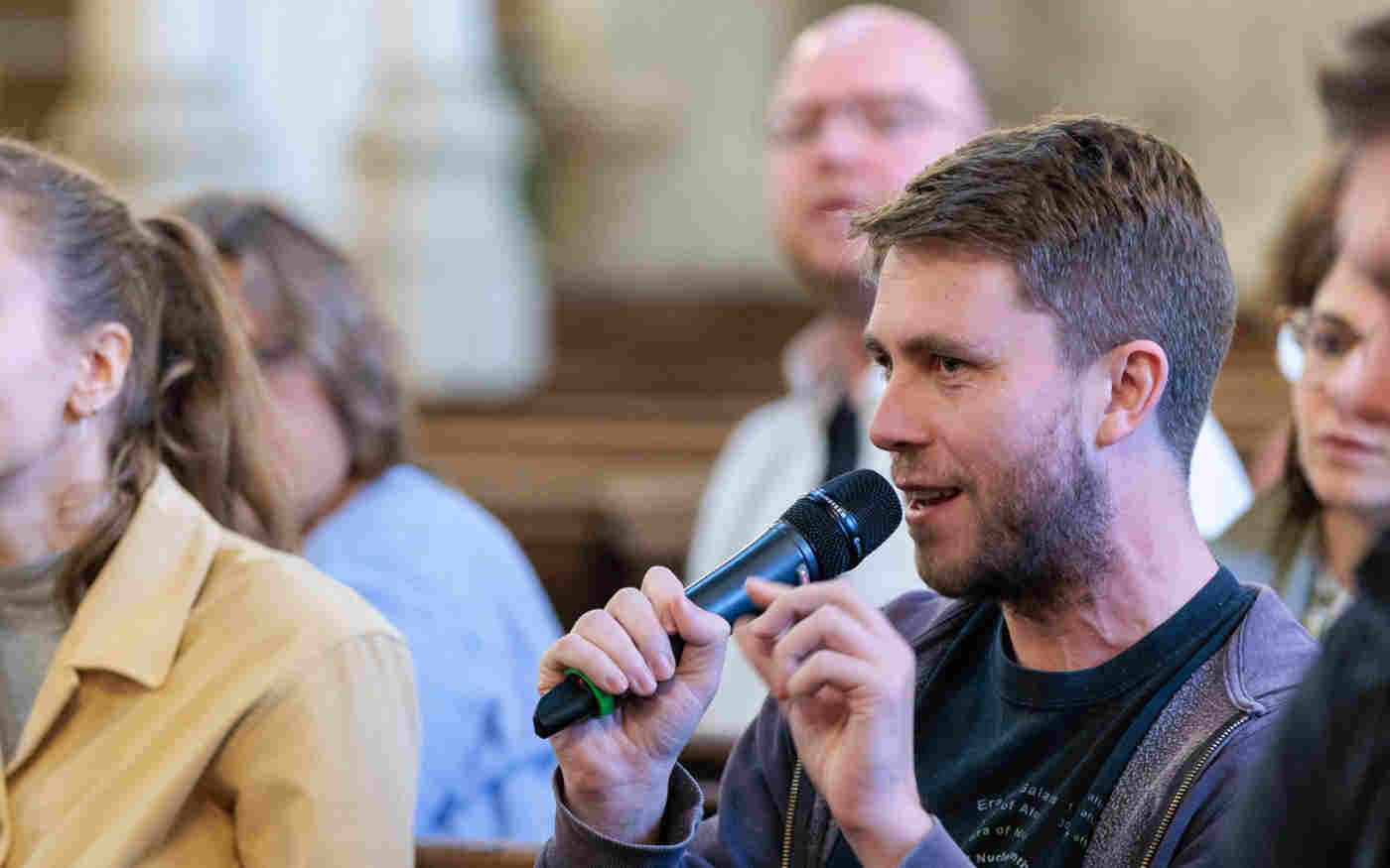 Photograph of an audience member holding a microphone asking a question surrounded by other people in St Margaret's Church