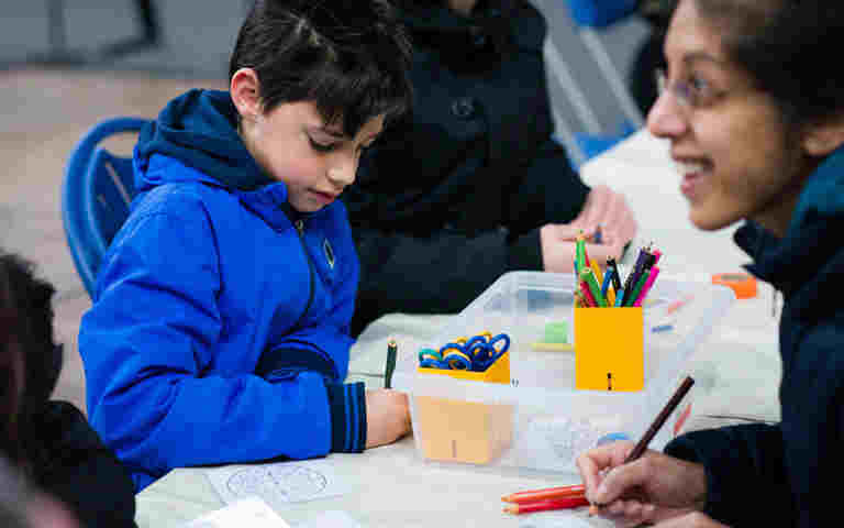 Photograph of a boy colouring in, surrounded by craft supplies on a table, with a female adult looking across the table, representing the activity pack for the first episode of Coronation Club